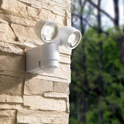 A white LED outdoor spotlight with a motion sensor mounted on a stone wall, with blurred green trees in the background.