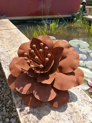 A rusty metal flower decoration sits on a stone ledge next to a pond with water lilies and plants.
