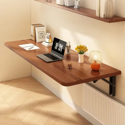 A compact wooden desk mounted on a wall above a radiator, featuring a laptop, notebook, and decorative items.