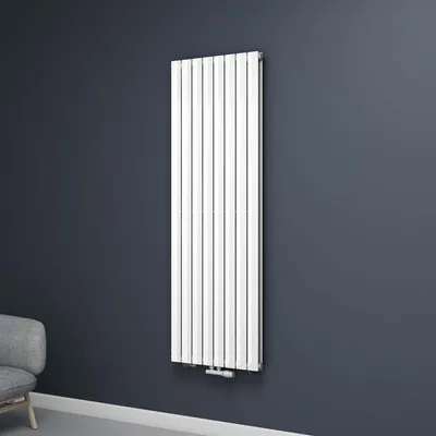 A white vertical radiator mounted on a dark gray wall, with a sliver of a gray armchair visible in the bottom left corner.