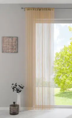 A sheer, sand-colored string curtain hangs in front of a large window, with a view of green trees and a bright sky outside. A decorative wooden plaque hangs on the wall to the left, and a vase with dark decorative branches sits on the floor.