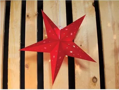 A red paper star lantern with small white stars printed on it, hanging in front of a wooden plank background.