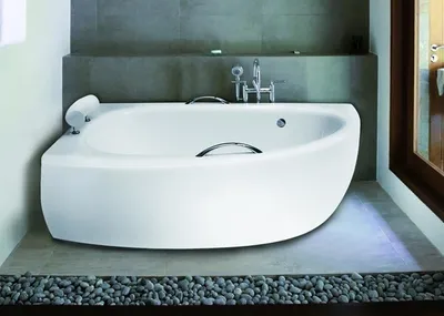 A modern white corner bathtub in a bathroom setting with grey tiled walls and a floor decorated with pebbles.