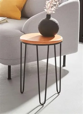 A small round side table with a wooden top and black metal hairpin legs, placed next to a gray sofa with a yellow cushion. A dark vase with dried flowers sits on top of the table.