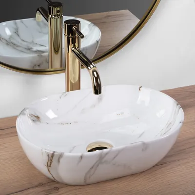 A close-up shot of a marble-patterned vessel sink with a gold faucet, placed on a wooden surface, with a mirror reflecting the sink and faucet in the background.