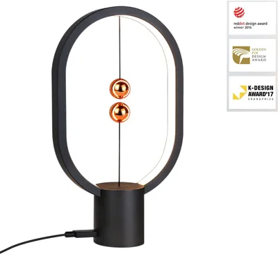 A black oval-shaped desk lamp with two copper spheres suspended in the middle by a thin wire, sitting on a black cylindrical base. The lamp has an LED light strip around the inside of the oval frame. There are award logos on the right side of the image.