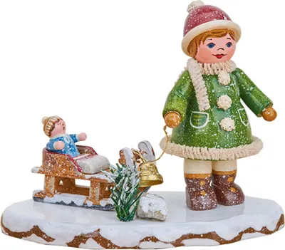 Ein fröhliches Winterkind in grüner Kleidung steht auf einer schneebedeckten Basis neben einem Schlitten mit einem sitzenden Kind und einem kleinen Weihnachtsbaum.
