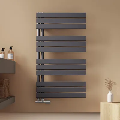 A modern anthracite towel radiator mounted on a light brown wall, with a small shelf and soap dispensers to the left, and a small wooden block with a vase of dried flowers to the right.