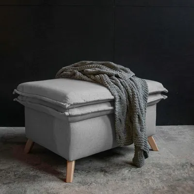 A gray ottoman with a knitted blanket draped over it, set against a dark textured wall and a concrete floor.