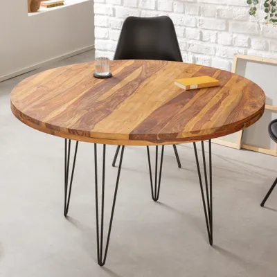 A round wooden dining table with black hairpin legs is shown in a room with a brick wall and a black chair.