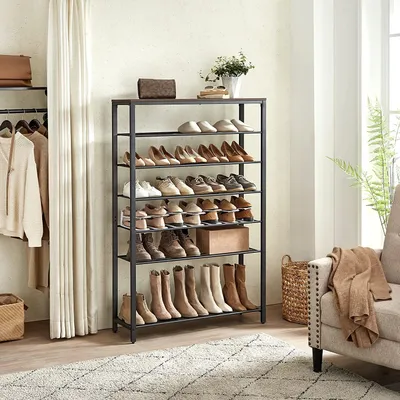 A multi-tiered shoe rack filled with shoes and boots, placed in a room with clothing on a rack to the left and a chair to the right.