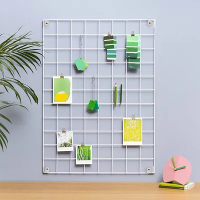 A white metal grid wall organizer with various items attached, including photos, color swatches, and pencils, displayed on a gray wall above a wooden desk with a pink clock.