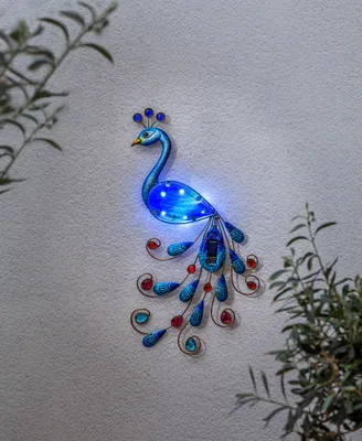 A decorative peacock solar light is mounted on a textured wall, with greenery on either side.