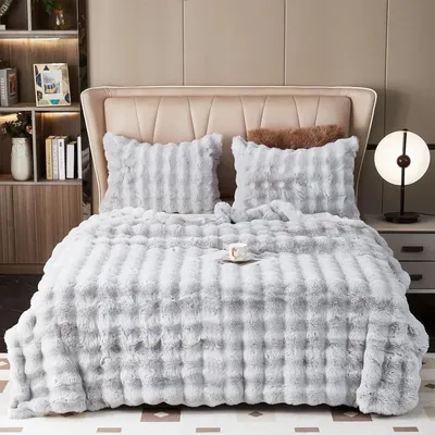 A plush blanket and pillows are neatly arranged on a bed, with a bedside table and lamp visible.