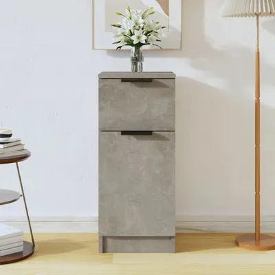 A tall, narrow grey cabinet with two drawers sits against a white wall, with a vase of white flowers on top. A floor lamp is to the right, and a small round table with books is to the left.