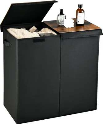 A black laundry hamper with two compartments and a wooden lid is shown. The lid is open, revealing towels inside one compartment. Two bottles and a white object are on the lid.