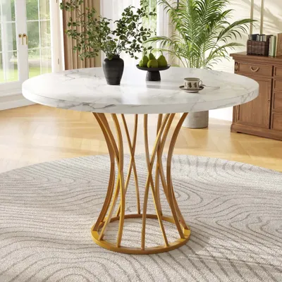A round marble-top dining table with a gold metal base is set in a well-lit dining room with large windows and plants.