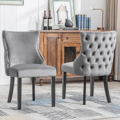 Two gray velvet dining chairs are shown in a room setting with a wooden cabinet in the background.