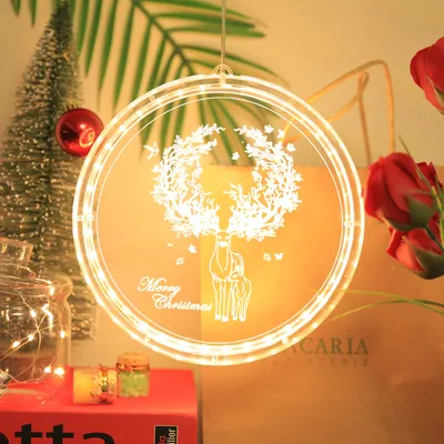 A circular LED Christmas decoration featuring a deer and wreath design, hanging in front of a blurred background with Christmas decorations.