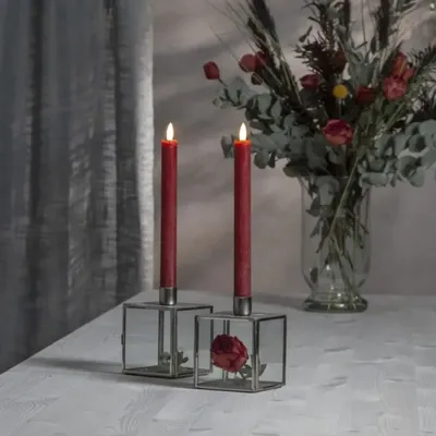 Two red LED taper candles in glass candle holders on a white table, with a floral arrangement in the background.