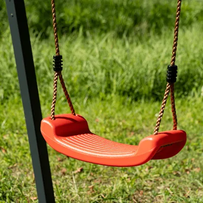 A red swing seat hangs from brown ropes attached to a dark gray swing set frame, set against a background of green grass.