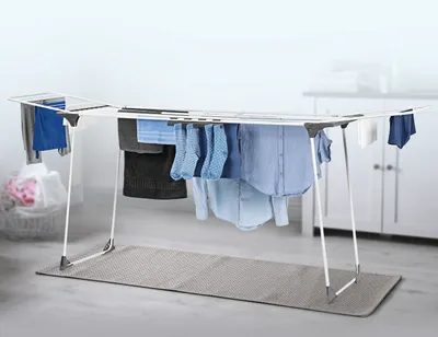 A white and gray extendable clothes drying rack with laundry hanging on it, placed on a gray mat in a room with a white cabinet in the background.