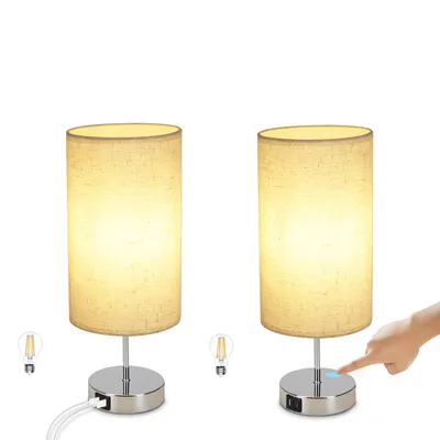 Two identical bedside lamps are shown against a white background. One lamp has a hand touching its base, indicating a touch feature. Small icons representing light bulbs are placed near the base of each lamp.