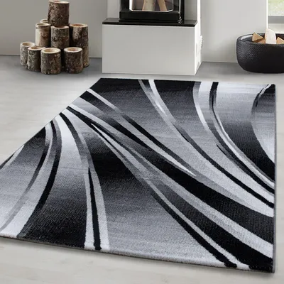 A modern abstract wave design rug in black, gray, and white is displayed on a light gray floor in a living room setting. In the background, there is a fireplace with logs and a basket of firewood.