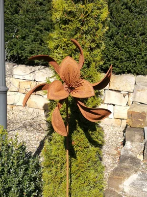 A rusty metal flower sculpture is placed in a garden setting, in front of a stone wall and green bushes.