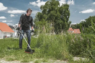 A man is using a string trimmer to cut tall grass in a yard with houses and trees in the background.
