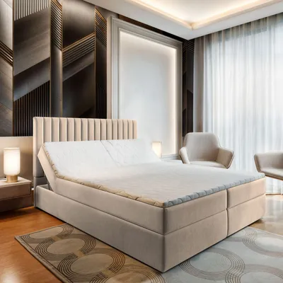 A bedroom scene featuring a large box spring bed with a beige upholstered headboard and base, set against a modern, textured wall. Two armchairs are visible in the background, along with sheer curtains.