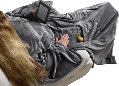 A person is lying on a couch, wrapped in a large gray blanket with sleeves, holding an e-reader.