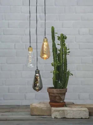 Three decorative LED light bulbs hang in front of a white brick wall, next to a potted cactus.