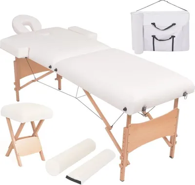 A white massage table with a headrest, armrests, and leg supports is displayed alongside a folded stool and two cylindrical bolsters. A white carrying bag is also shown in the upper right corner.