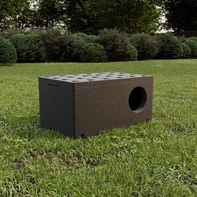 A dark brown insulated cat house sits on a grassy lawn with bushes and trees in the background.
