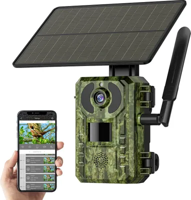 A hand holds a smartphone displaying a live feed from a trail camera, which is also shown in the image. The trail camera is mounted on a solar panel.
