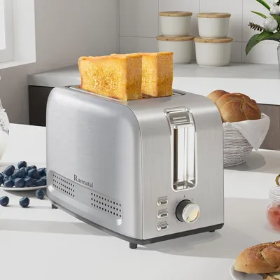 A silver toaster with two slices of toast in it, placed on a white countertop with a bowl of blueberries and some bread rolls in a basket in the background.