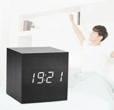 A man is stretching in bed next to a digital alarm clock.