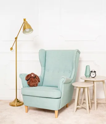 A plush bunny sits on a teal armchair next to two small side tables, one with a vase and alarm clock, and a floor lamp stands to the left.
