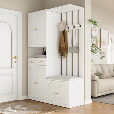 A white entryway storage set with a bench, coat hooks, and cabinets, placed next to a white door and in front of a living room with a sofa.