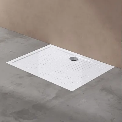 A white rectangular shower tray with a silver drain is placed on a concrete floor against a brown wall.