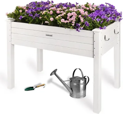 A white raised garden bed filled with purple and pink flowers, with a watering can and trowel placed on the ground below.