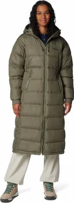 A woman wearing a long green puffer coat and light-colored pants, standing against a white background.