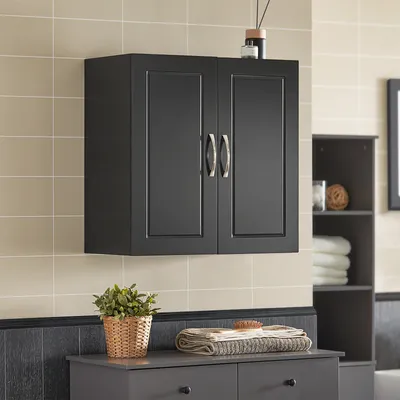A black wall-mounted cabinet with two doors is shown against a tiled wall. Below the cabinet is a gray dresser with a potted plant and towels on top. A dark gray shelving unit is visible to the right.