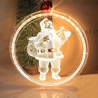 A close-up of a lit-up Christmas ornament featuring Santa Claus, with a small Christmas tree in the background.