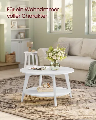 A white round coffee table with a shelf is placed on a patterned rug in a living room. A vase of flowers sits on the table, and a sofa is visible in the background.