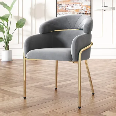 A grey velvet armchair with gold metal legs is shown in a room with wood flooring and a plant.