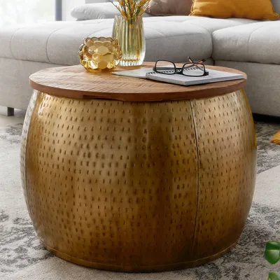 A round coffee table with a hammered gold metal base and a wooden top, placed in a living room setting with a sofa and rug.