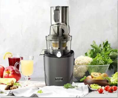A Kuvings REVO830 Slow Juicer is displayed prominently on a table, surrounded by fresh fruits and vegetables, with glasses of juice nearby.