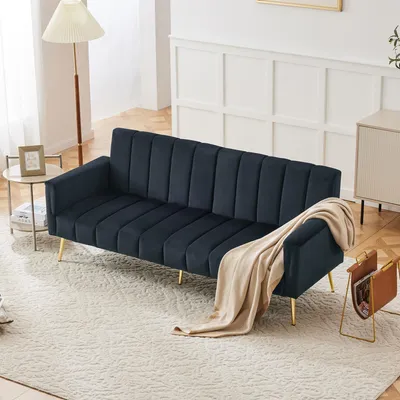 A dark blue sofa with gold legs is placed in a well-lit living room, with a beige throw draped over the armrest. A floor lamp and a side table with a picture frame are visible to the left, and a magazine rack is to the right.
